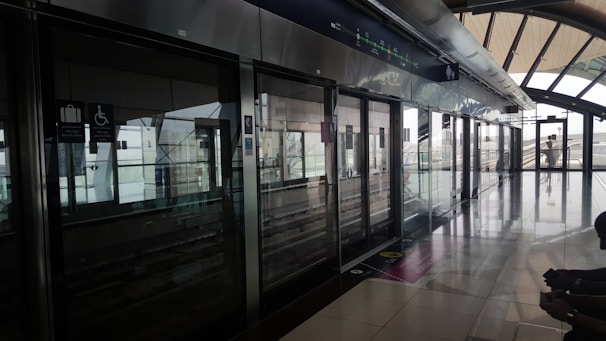 A modern, clean station platform with sleek metallic finishing and large glass windows. Signs for luggage and accessibility features are visible above sliding doors. The platform is empty, emphasizing a quiet and orderly environment with natural light streaming in.