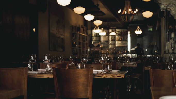 A dimly lit restaurant with wooden tables and chairs. Each table is set with clean glassware and white napkins. The ambient lighting from chandeliers and wall-mounted fixtures creates a warm, cozy atmosphere. In the background, a bar area with shelves stocked with bottles and a person in silhouette can be seen.