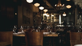 A dimly lit restaurant with wooden tables and chairs. Each table is set with clean glassware and white napkins. The ambient lighting from chandeliers and wall-mounted fixtures creates a warm, cozy atmosphere. In the background, a bar area with shelves stocked with bottles and a person in silhouette can be seen.