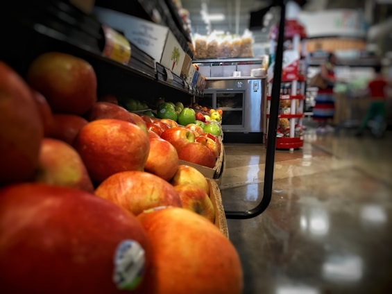 A grocery store aisle with a close-up perspective of red and green apples displayed on a low shelf. In the background, there are other produce shelves and some aisles with various products visible. The image has a shallow depth of field, focusing on the apples in the foreground while the background appears blurred. The floor is glossy, reflecting the surroundings.