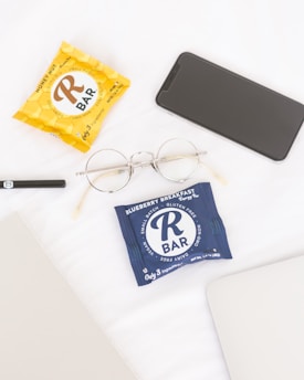 On a white surface, several items are neatly arranged including two packaged energy bars, one in a yellow wrapper labeled 'Honey Nut' and another in a blue wrapper labeled 'Blueberry Breakfast.' Nearby are gold-rimmed glasses, a black smartphone, an open gray laptop, and a black pen.