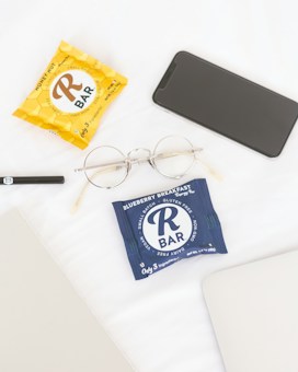 On a white surface, several items are neatly arranged including two packaged energy bars, one in a yellow wrapper labeled 'Honey Nut' and another in a blue wrapper labeled 'Blueberry Breakfast.' Nearby are gold-rimmed glasses, a black smartphone, an open gray laptop, and a black pen.