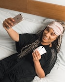 A person lies comfortably on a bed, wearing a pink sleep mask pushed up on their forehead and holding a smartphone in their outstretched hand. They are smiling and appear to be taking a selfie. In the other hand, they hold two small tubes, likely cosmetic products. The bedding is white, and the person is dressed in a black garment.
