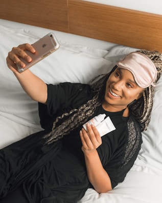 A person lies comfortably on a bed, wearing a pink sleep mask pushed up on their forehead and holding a smartphone in their outstretched hand. They are smiling and appear to be taking a selfie. In the other hand, they hold two small tubes, likely cosmetic products. The bedding is white, and the person is dressed in a black garment.