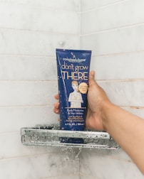 A hand holding a dark blue tube of body moisturizer and hair inhibitor in a shower setting. The tube has text and an illustration of a person on it, and water droplets are visibly splashing on both the tube and the white tiled shower wall.