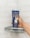 A hand holding a dark blue tube of body moisturizer and hair inhibitor in a shower setting. The tube has text and an illustration of a person on it, and water droplets are visibly splashing on both the tube and the white tiled shower wall.