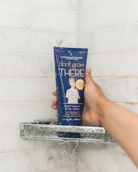 A hand holding a dark blue tube of body moisturizer and hair inhibitor in a shower setting. The tube has text and an illustration of a person on it, and water droplets are visibly splashing on both the tube and the white tiled shower wall.