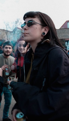 A group of young people are gathered outdoors, engaging in casual conversation. One person in the foreground is wearing sunglasses, has a cigarette, and is holding a drink. The others in the background also appear to be holding drinks and are dressed casually.