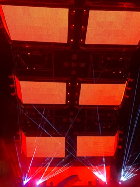 Large red LED screens mounted on a dark ceiling, surrounded by beams of blue and red lights, creating a vibrant and dynamic atmosphere.