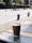 A brown paper cup with a percentage symbol on it is sitting on a tiled table. The background features an outdoor setting with a pavement, green bushes, and some blurred people walking.