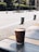 A brown paper cup with a percentage symbol on it is sitting on a tiled table. The background features an outdoor setting with a pavement, green bushes, and some blurred people walking.