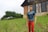 A plastic male doll dressed in a blue t-shirt and red pants stands on a grassy field in front of a rustic house. The house has large windows with white curtains, stone accents, and a wooden upper section. In the background, there are green trees against a cloudy sky.
