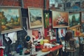 A collection of framed paintings and various items displayed in an open market setting. The paintings feature landscapes and floral arrangements. Other objects, such as ceramic pieces, mirrors, and bottles, are placed on tables. There's also a mannequin head displayed.