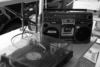 A vintage boombox and a record player with a transparent dust cover are placed on a table. Several cables are visible connecting the devices, and there are boxes in the background.