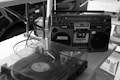 A vintage boombox and a record player with a transparent dust cover are placed on a table. Several cables are visible connecting the devices, and there are boxes in the background.