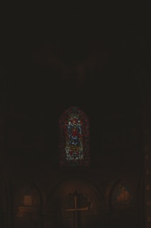 A dimly lit interior of a church featuring a stained glass window depicting a religious figure, likely Jesus, with rich colors and intricate designs. Below the window, a large wooden cross stands prominently. The overall atmosphere is solemn and reverent, consistent with a place of worship.