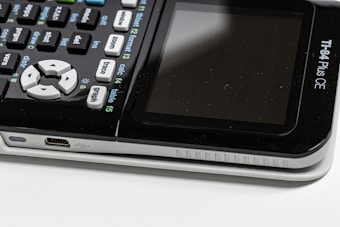 Close-up view of a TI-84 Plus CE calculator with a focus on the keypad and the side USB port. The display screen is visible but turned off, and the calculator shows various buttons with mathematical functions.