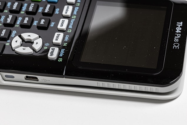 Close-up view of a TI-84 Plus CE calculator with a focus on the keypad and the side USB port. The display screen is visible but turned off, and the calculator shows various buttons with mathematical functions.