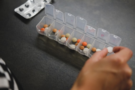 A hand is organizing various pills into a transparent plastic pill organizer with individual compartments, each marked for different days or times. There is also a blister pack in the background.