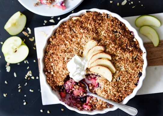 A baked dessert with a golden-brown crumble topping is served in a round, white dish. Slices of fresh apple and a dollop of cream are placed on top. A spoon is inserted into the dessert, revealing a colorful filling of fruit underneath. Sliced apples and crumbs are scattered around on a dark surface.