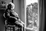 An elderly woman sits in a well-lit room by a window, looking contemplatively outside. She wears a necklace and a cardigan. The room includes a framed photograph on the wall and there are plants on a balcony visible through the window.