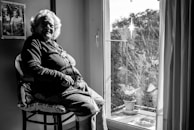 An elderly woman sits in a well-lit room by a window, looking contemplatively outside. She wears a necklace and a cardigan. The room includes a framed photograph on the wall and there are plants on a balcony visible through the window.
