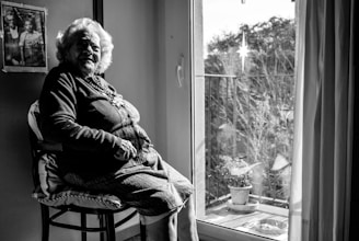 An elderly woman sits in a well-lit room by a window, looking contemplatively outside. She wears a necklace and a cardigan. The room includes a framed photograph on the wall and there are plants on a balcony visible through the window.