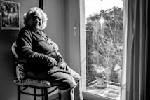 An elderly woman sits in a well-lit room by a window, looking contemplatively outside. She wears a necklace and a cardigan. The room includes a framed photograph on the wall and there are plants on a balcony visible through the window.
