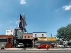 A city street scene showing an auto repair shop and tire shop with large signboards. There is a bright orange sports car parked in front of the shop, with another vehicle partially visible inside the garage. The building housing the shop is made of brick, and there is a large, empty billboard structure above it. To the right, some trees and different buildings are visible under a clear blue sky.