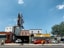 A city street scene showing an auto repair shop and tire shop with large signboards. There is a bright orange sports car parked in front of the shop, with another vehicle partially visible inside the garage. The building housing the shop is made of brick, and there is a large, empty billboard structure above it. To the right, some trees and different buildings are visible under a clear blue sky.