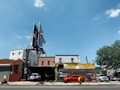 A city street scene showing an auto repair shop and tire shop with large signboards. There is a bright orange sports car parked in front of the shop, with another vehicle partially visible inside the garage. The building housing the shop is made of brick, and there is a large, empty billboard structure above it. To the right, some trees and different buildings are visible under a clear blue sky.