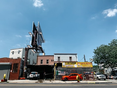 A city street scene showing an auto repair shop and tire shop with large signboards. There is a bright orange sports car parked in front of the shop, with another vehicle partially visible inside the garage. The building housing the shop is made of brick, and there is a large, empty billboard structure above it. To the right, some trees and different buildings are visible under a clear blue sky.