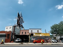 A city street scene showing an auto repair shop and tire shop with large signboards. There is a bright orange sports car parked in front of the shop, with another vehicle partially visible inside the garage. The building housing the shop is made of brick, and there is a large, empty billboard structure above it. To the right, some trees and different buildings are visible under a clear blue sky.