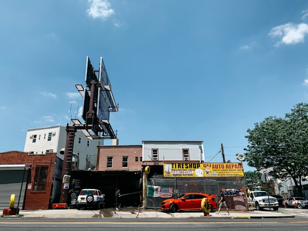A city street scene showing an auto repair shop and tire shop with large signboards. There is a bright orange sports car parked in front of the shop, with another vehicle partially visible inside the garage. The building housing the shop is made of brick, and there is a large, empty billboard structure above it. To the right, some trees and different buildings are visible under a clear blue sky.