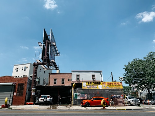 A city street scene showing an auto repair shop and tire shop with large signboards. There is a bright orange sports car parked in front of the shop, with another vehicle partially visible inside the garage. The building housing the shop is made of brick, and there is a large, empty billboard structure above it. To the right, some trees and different buildings are visible under a clear blue sky.