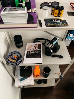 A shelving unit contains various personal items including a stack of Polaroid photos, a film camera, sunglasses, a 'Save the Date' card, and film rolls on the top shelf. The middle shelf has a can of butter cookies, a book or magazine titled 'RUNAWAY', a black mug, and a closed pocket knife. The bottom shelf holds a New York Yankees cap, an orange water bottle, and some mail.