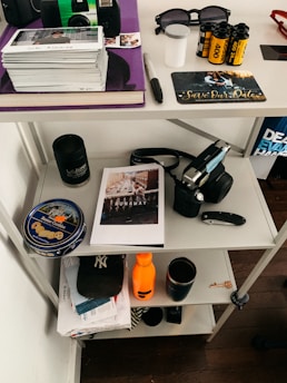 A shelving unit contains various personal items including a stack of Polaroid photos, a film camera, sunglasses, a 'Save the Date' card, and film rolls on the top shelf. The middle shelf has a can of butter cookies, a book or magazine titled 'RUNAWAY', a black mug, and a closed pocket knife. The bottom shelf holds a New York Yankees cap, an orange water bottle, and some mail.