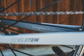 A close-up of a bicycle frame featuring the text 'Created in Germany'. The frame is partially obscured by a tilted angle, with a chain and a blurry wooden background.