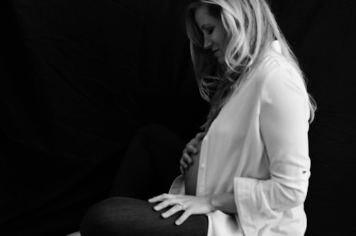 A black and white portrait of a woman seated against a dark background. She is wearing a light-colored shirt, and her long hair falls over her shoulders. Her hand rests gently on her belly, suggesting a contemplative and serene moment.