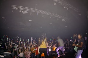 A band is performing on stage in front of an energetic crowd. The audience is holding colorful glow sticks and cheering. The stage is equipped with lighting and sound equipment, and the band members are engaged in their performance with instruments like a guitar.