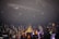 A band is performing on stage in front of an energetic crowd. The audience is holding colorful glow sticks and cheering. The stage is equipped with lighting and sound equipment, and the band members are engaged in their performance with instruments like a guitar.