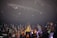 A band is performing on stage in front of an energetic crowd. The audience is holding colorful glow sticks and cheering. The stage is equipped with lighting and sound equipment, and the band members are engaged in their performance with instruments like a guitar.
