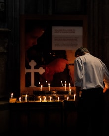 A person in a white shirt is seen lighting candles in a dimly lit setting, next to a sign welcoming people to light a candle. The area is illuminated by the glow of numerous candles placed on a stand. A cross is visible on a board with a background showing a painting or photo featuring warm colors.