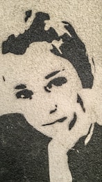 A monochrome stencil art of a face on a textured wall, showing prominent features with black paint against a beige background.