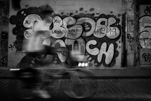 Graffiti-covered wall with bold and abstract designs in large letters. A motion-blurred figure on a bicycle passes in front of the wall, adding a sense of movement to the scene. The environment appears urban, with brick paving on the ground.