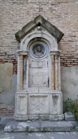 A sculptural, ornate stonework monument embedded into a large, weathered brick wall. The monument features a detailed arch with a peaked roof, supported by two slender columns. Within the arch is a circular medallion enclosing a carved portrait of a man's head. The structure shows signs of aging and has a cracked and worn appearance. There are some green plants growing at the base of the monument.