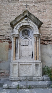A sculptural, ornate stonework monument embedded into a large, weathered brick wall. The monument features a detailed arch with a peaked roof, supported by two slender columns. Within the arch is a circular medallion enclosing a carved portrait of a man's head. The structure shows signs of aging and has a cracked and worn appearance. There are some green plants growing at the base of the monument.