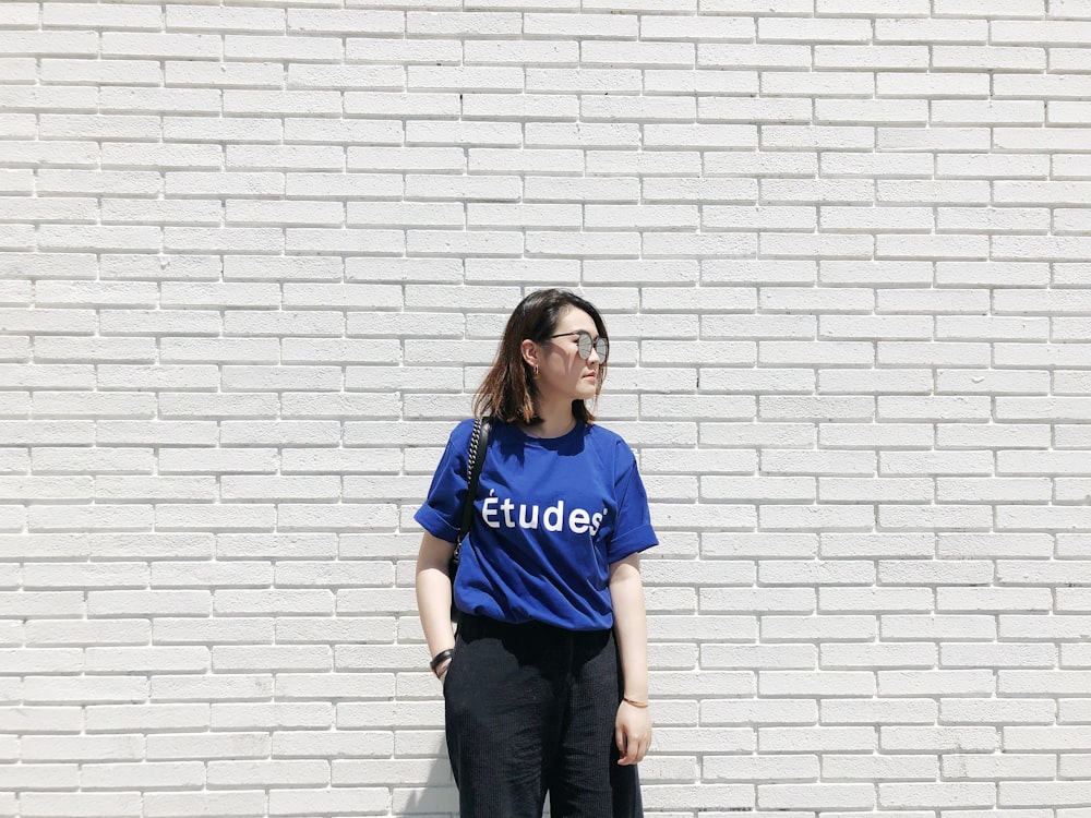 woman in blue Etudes shirt standing by the white brick wall