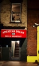 A building facade features a red sign with white lettering reading 'AMERICAN DREAM 168 BED & BREAKFAST' above a door with a glass panel revealing interior stairs. A window on the upper floor shows part of an industrial-looking room with a person visible, creating a scene suggestive of urban life. The walls are made of brick and painted surfaces, and there is a security camera mounted on the wall.