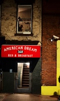 A building facade features a red sign with white lettering reading 'AMERICAN DREAM 168 BED & BREAKFAST' above a door with a glass panel revealing interior stairs. A window on the upper floor shows part of an industrial-looking room with a person visible, creating a scene suggestive of urban life. The walls are made of brick and painted surfaces, and there is a security camera mounted on the wall.